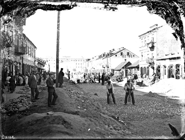 Fragment fotografii archiwalnej z przedstawieniem osób pracujących na ulicy. W tle zabudowa miejska (Siedlce)