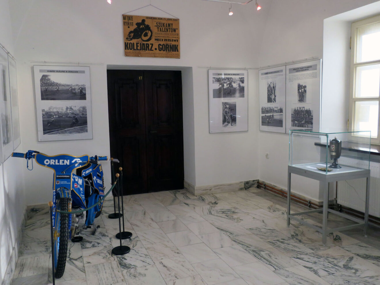 Fragmnet ekspozycji z planszami, plakatem meczu żużlowego, motocyklem wyścigowym i pamiątkowym trofeum w gablocie