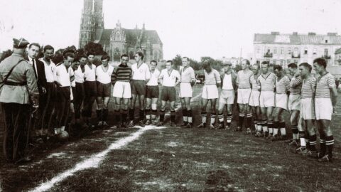 Fotografia archiwalna z przedstawieniem piłkarzy stojących w półkolu na środku boiska. W tle zabudowania miejskie.